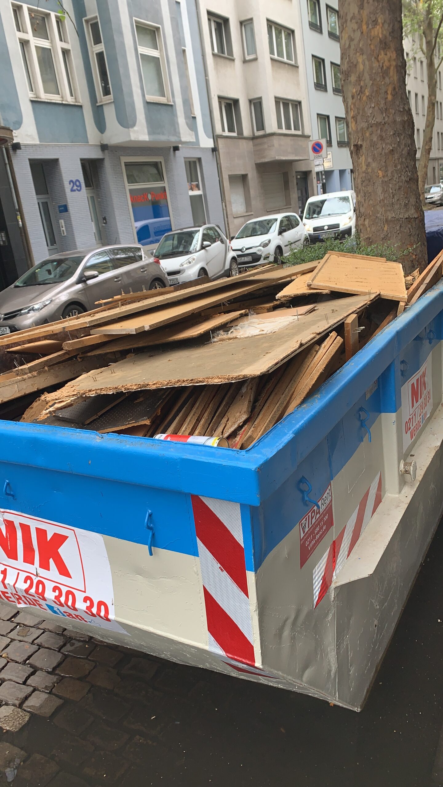 Großer Müllcontainer mit Holz- und Bauschutt steht auf einer Stadtstraße neben parkenden Autos und Gebäuden.