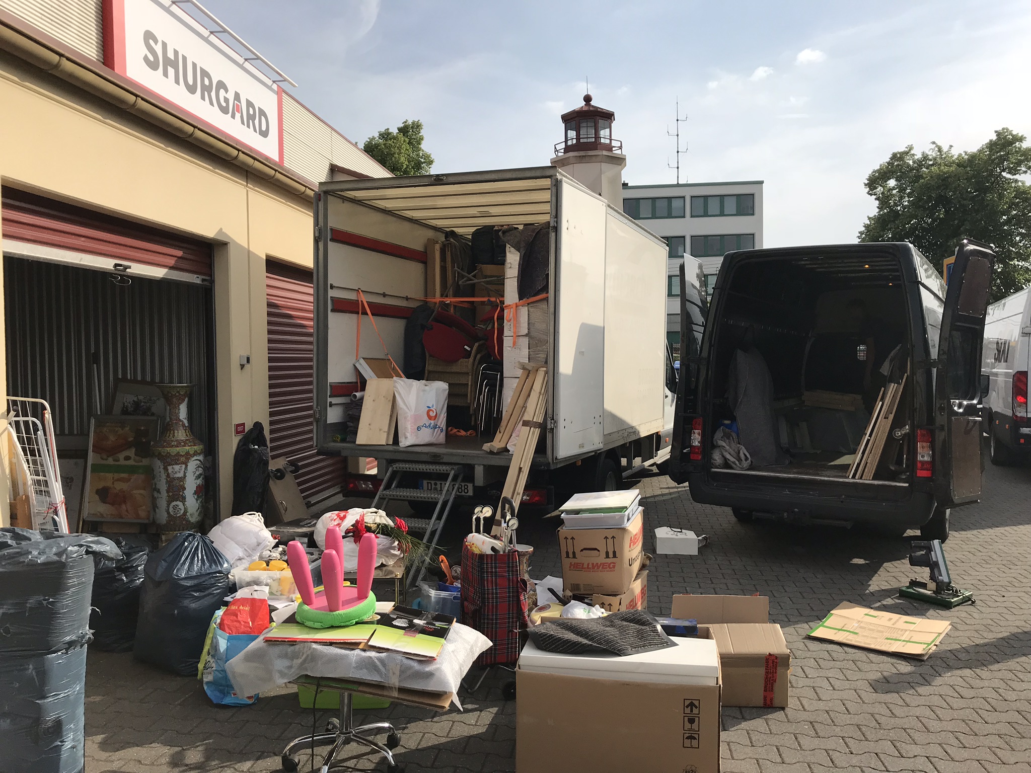 Umzugswagen und Transporter vor Lagerhalle, am Boden liegen verschiedene Kisten und Gegenstände.