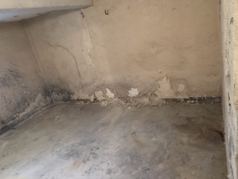 Abgeplatzte und rissige Farbe mit Wasserschäden an der unteren Wandecke eines Raumes mit Betonboden.