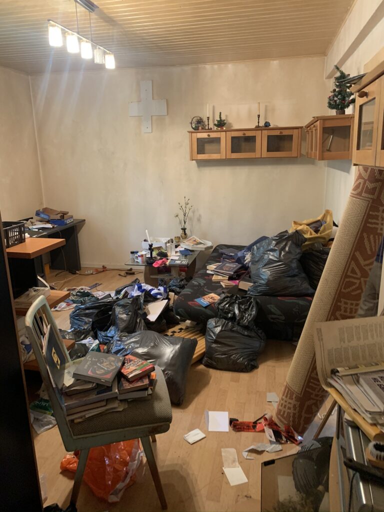 Unordentliches Zimmer mit verstreuten Papieren, Büchern, schwarzen Müllsäcken und durcheinanderstehenden Möbeln.