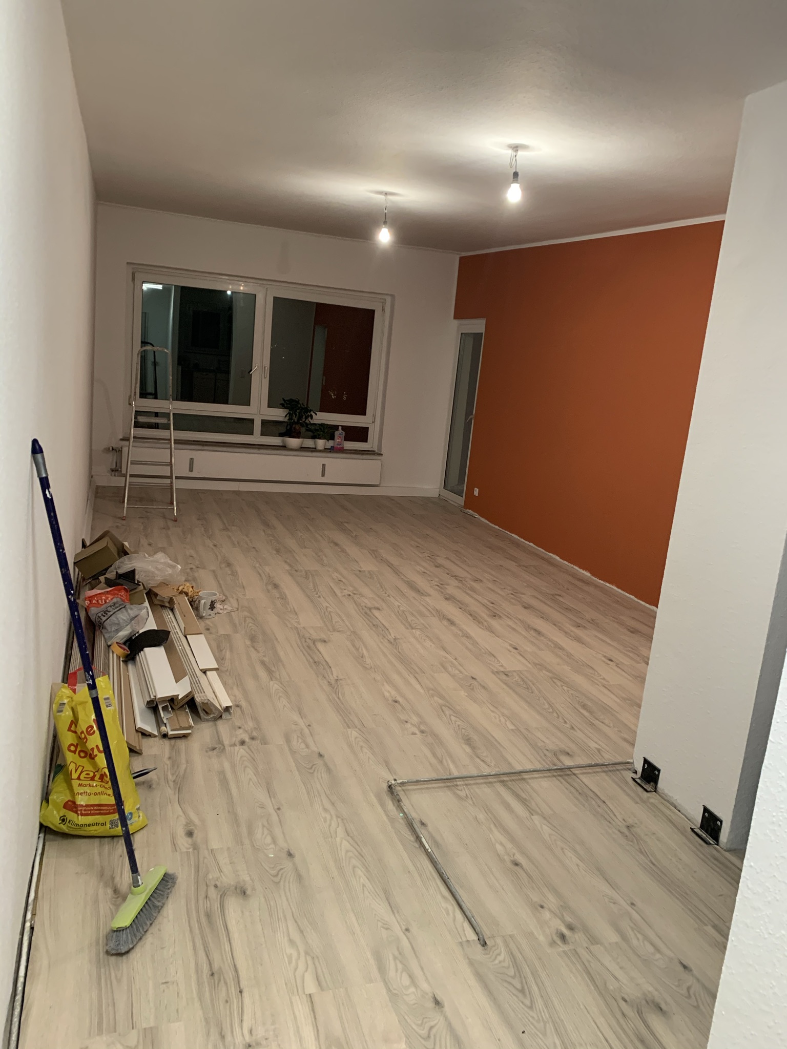 Handwerker renovieren einen Raum mit hellem Holzfußboden und oranger Wand, Werkzeug und Kartons liegen verstreut, Licht brennt.