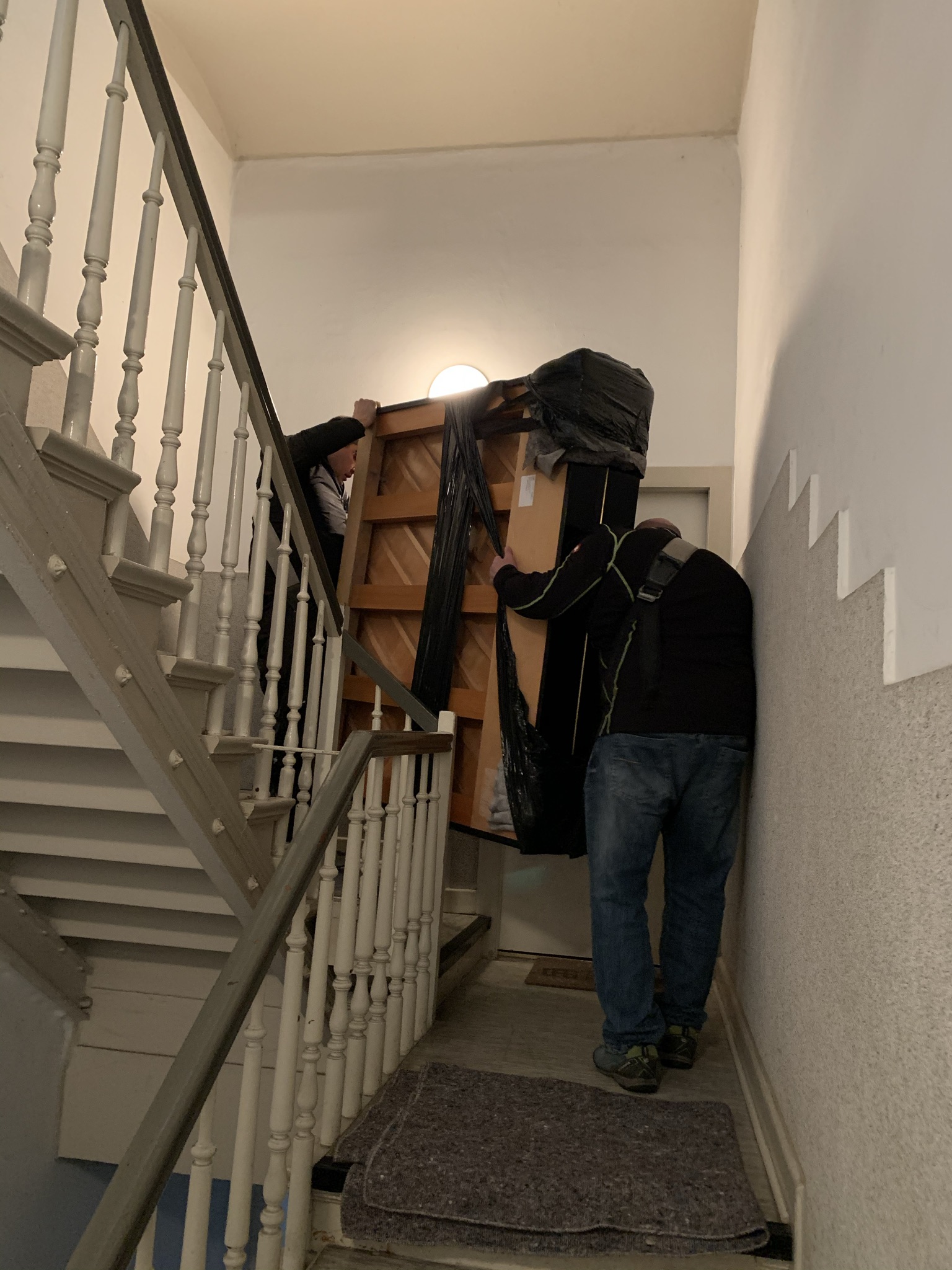 Zwei Personen tragen ein großes, verpacktes Möbelstück eine schmale Treppe mit weißen Geländern und beigen Wänden hinauf.